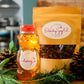 An image featuring a paper package labeled 'Elderberry Syrup Kit' with a clear window showing contents inside, alongside an 8 oz jar of honey with a red twist cap, both presented on a wooden surface with pine cones and greenery in the background.
